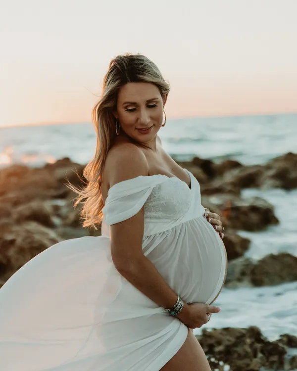 Expectant mother in a flowing white dress on a rocky shoreline at sunset