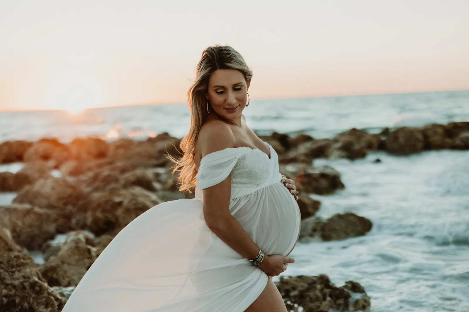 Expectant mother in a flowing white dress on a rocky shoreline at sunset