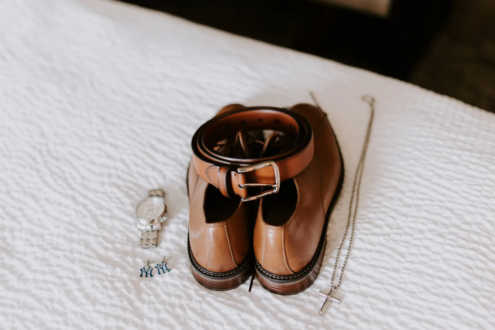 Groom's brown shoes, belt, watch and cross necklace laid out on a bed