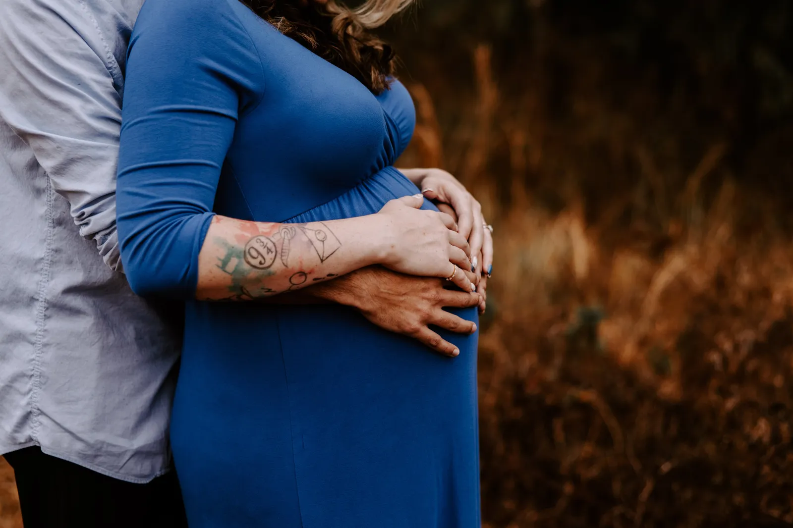 Close-up of his arms around her bump, her tattooed forearm resting over his