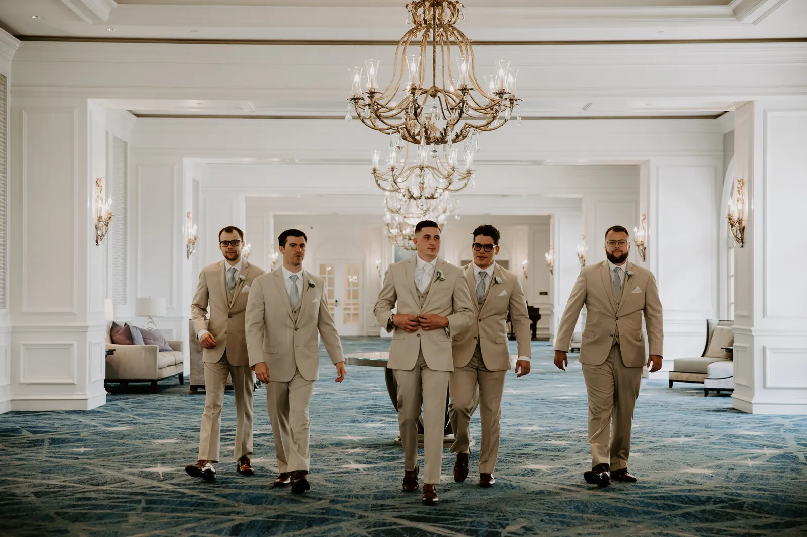 Groom and groomsmen in tan suits walking through a chandelier-lit hall at the Ritz-Carlton, Sarasota