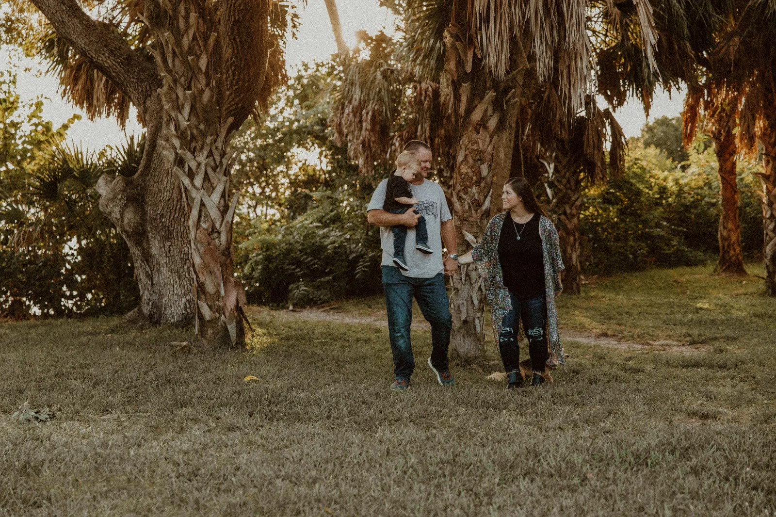 Family walking beneath palms and oaks, the father carrying their son