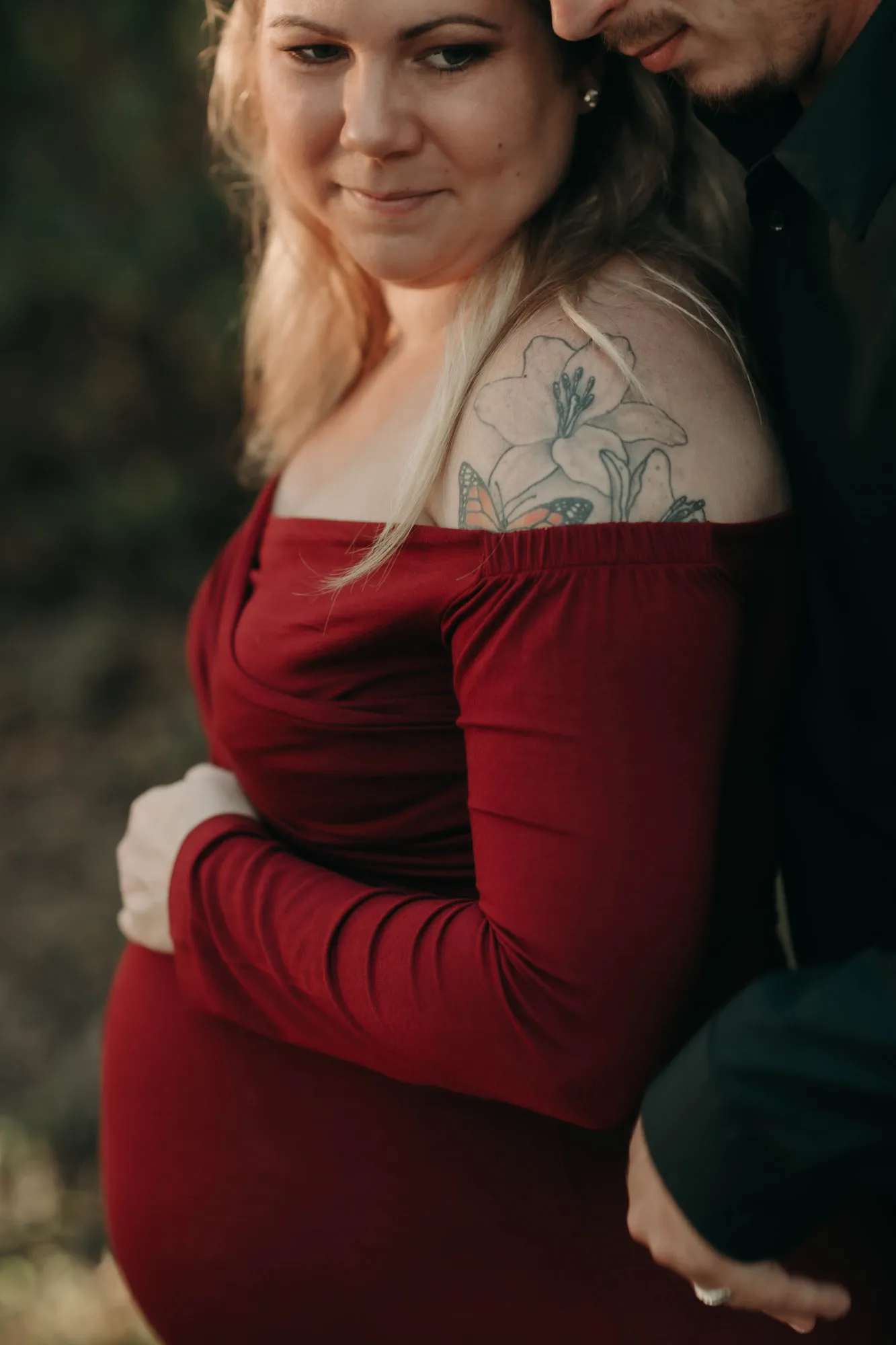 Expectant mother in an off-shoulder red dress, a floral tattoo on her shoulder, her partner behind her