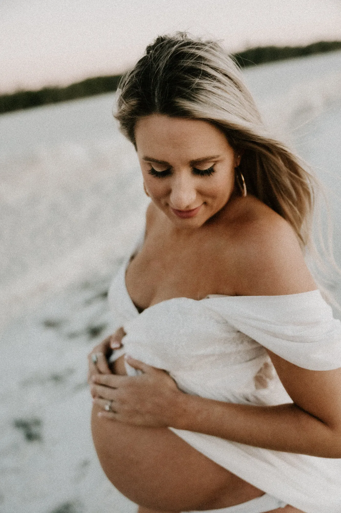 Expectant mother looking down, wrapped in white fabric on the sand