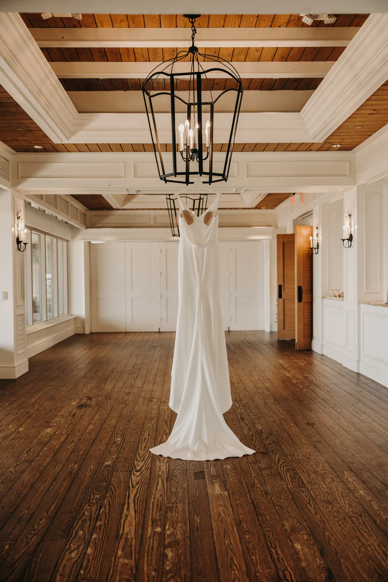 Wedding dress hanging from a lantern chandelier in a white room with wooden floors