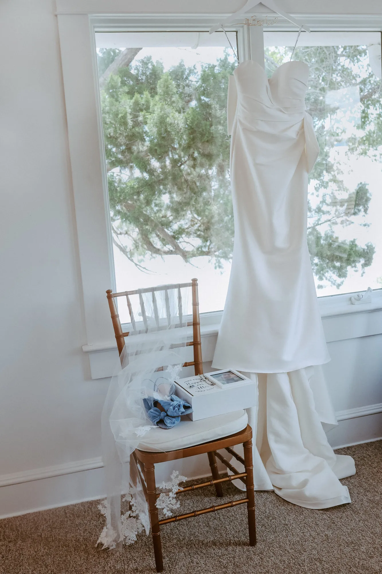 Wedding gown hanging in a sunlit window above a chair holding blue shoes