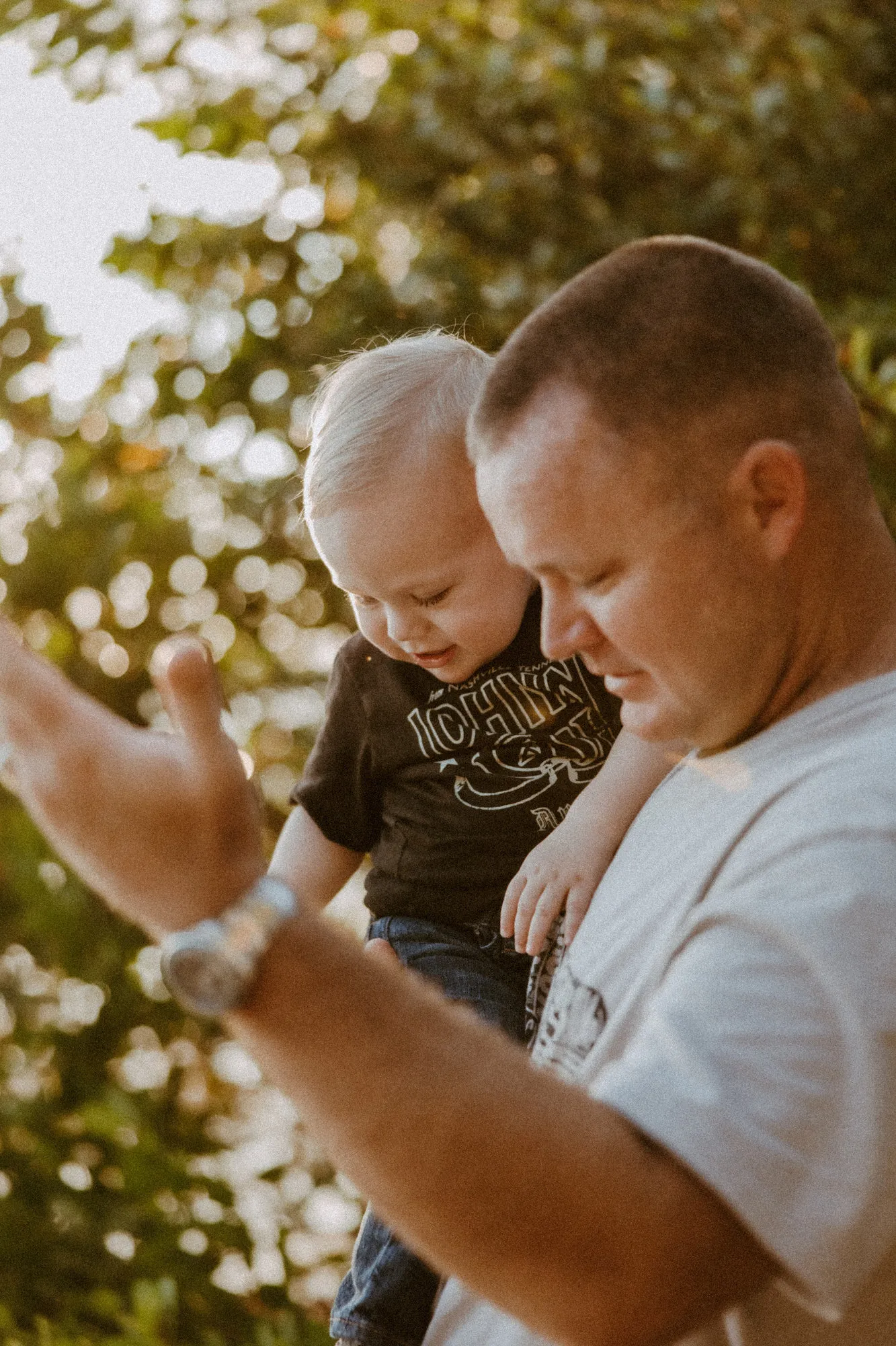 Father holding his toddler son, both looking down, as a hand waves in the foreground