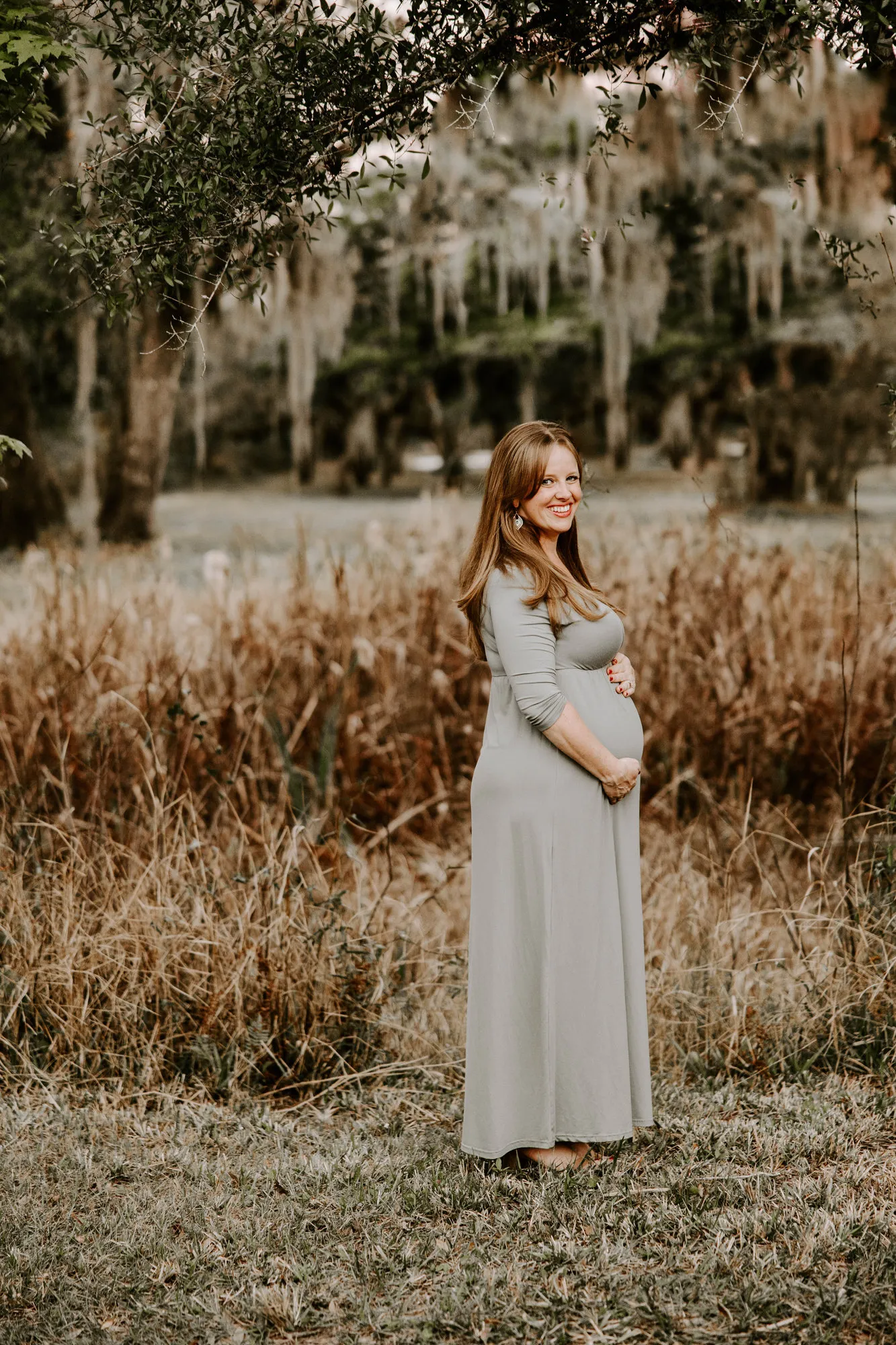 Expectant mother looking away and smiling, moss-draped oaks behind her