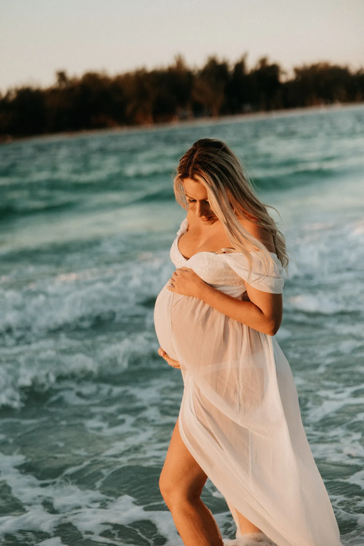 Expectant mother in a sheer white dress standing in the surf
