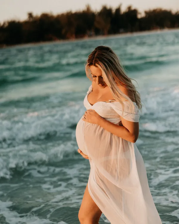 Expectant mother in a sheer white dress standing in the surf