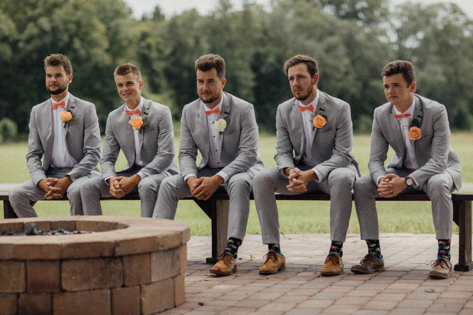 Five groomsmen in grey suits and coral bow ties sitting on a bench