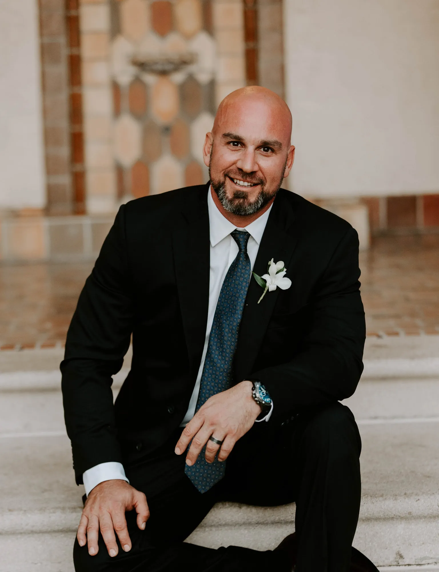 Groom in a dark suit and white boutonniere sitting on the steps, smiling