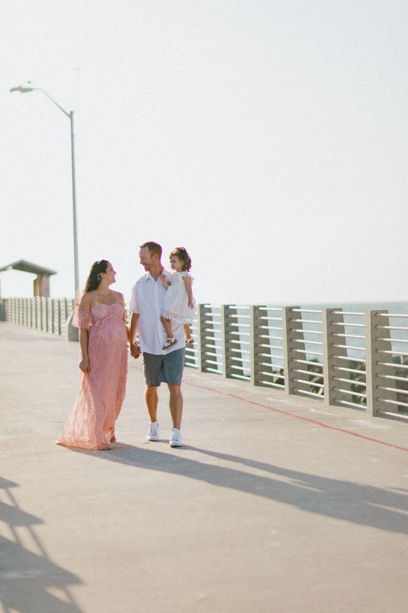 Expectant mother in a pink gown walking the pier beside her husband, who carries their daughter