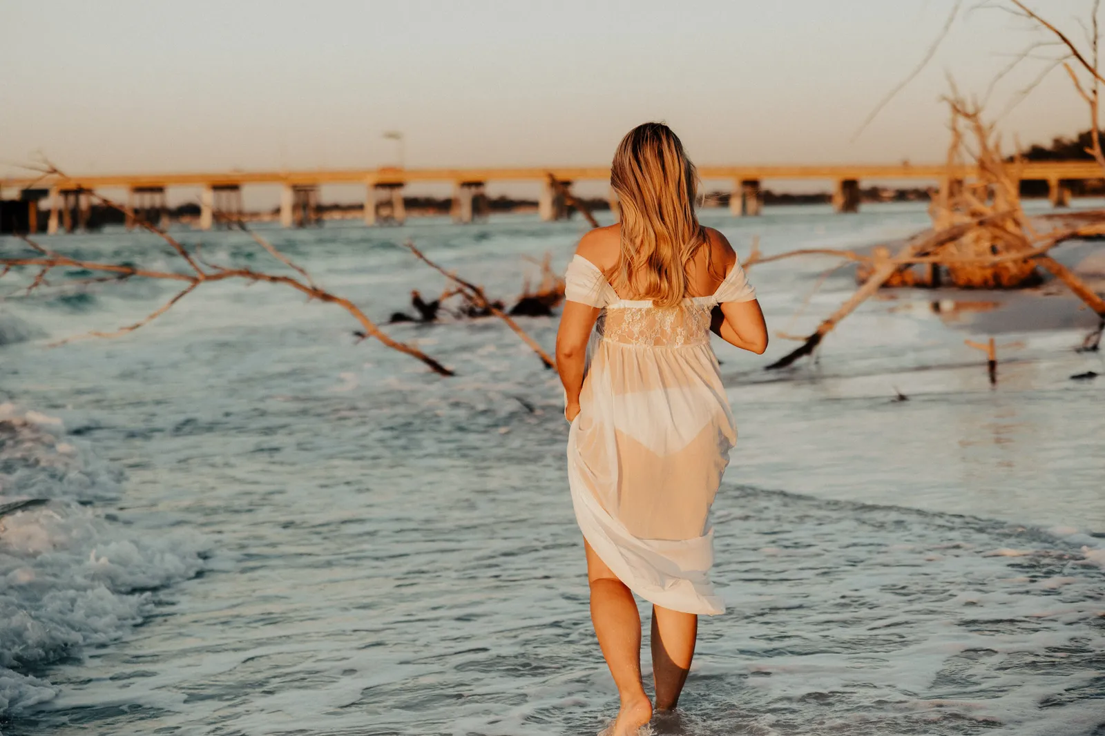 Expectant mother wading toward a pier and driftwood, seen from behind