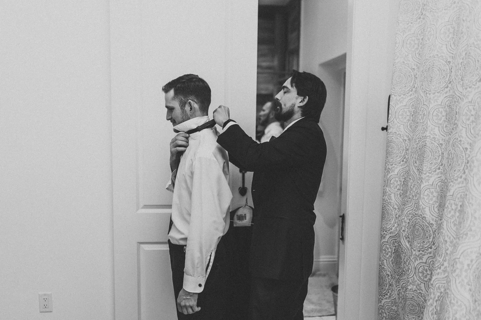 One man adjusting another's collar as they get dressed, in black and white