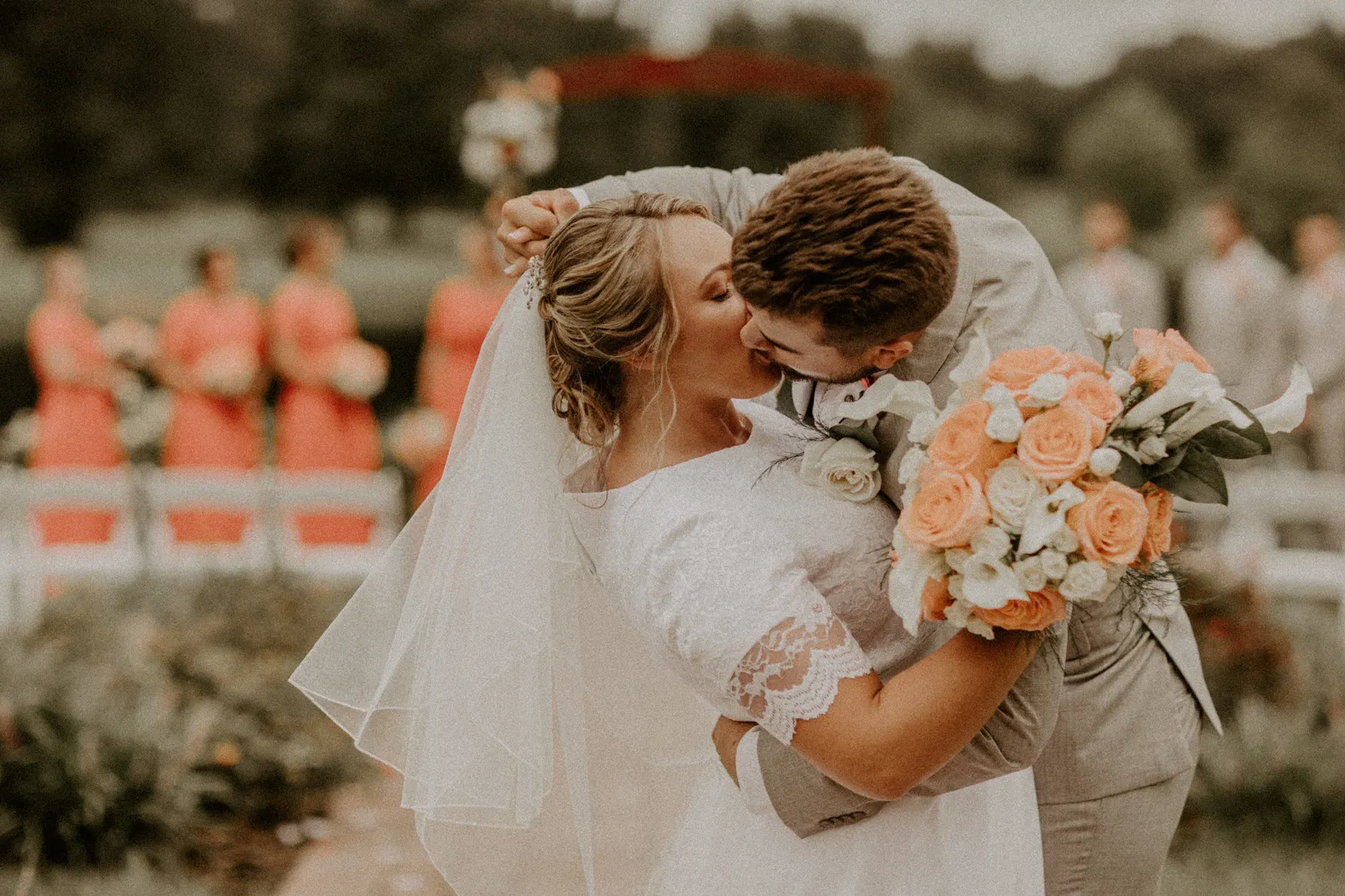 Groom kissing his bride as she holds a coral and peach bouquet