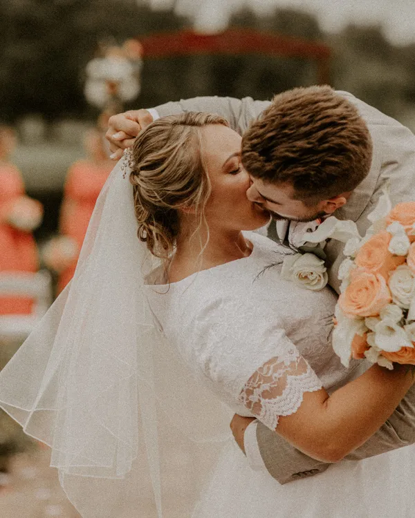Groom kissing his bride as she holds a coral and peach bouquet
