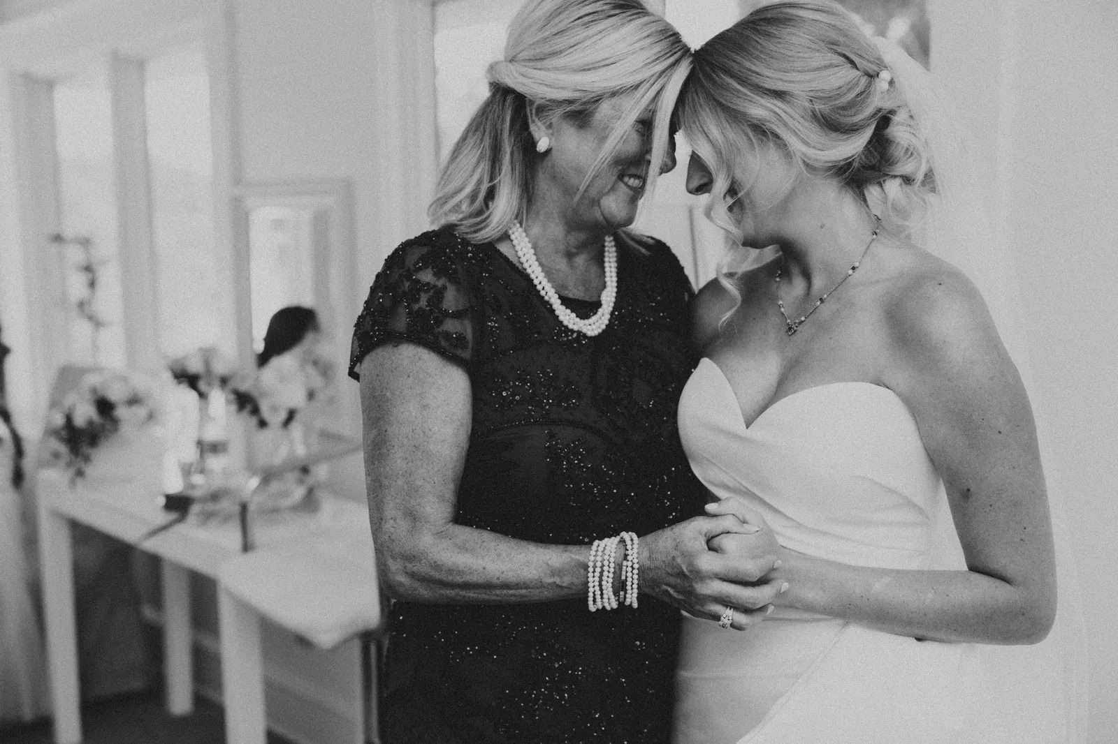 Bride and a woman in pearls touching foreheads, in black and white