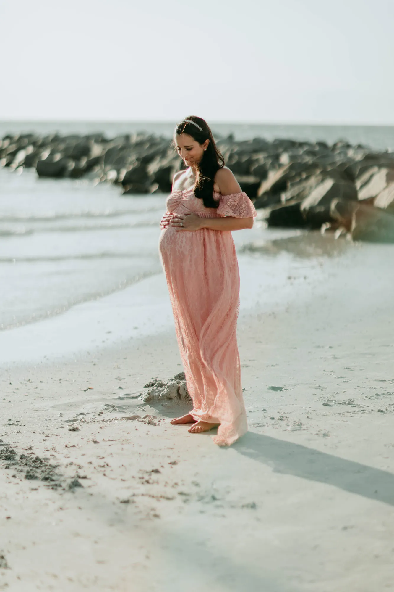 Expectant mother in a pink lace gown on the beach beside a rock jetty