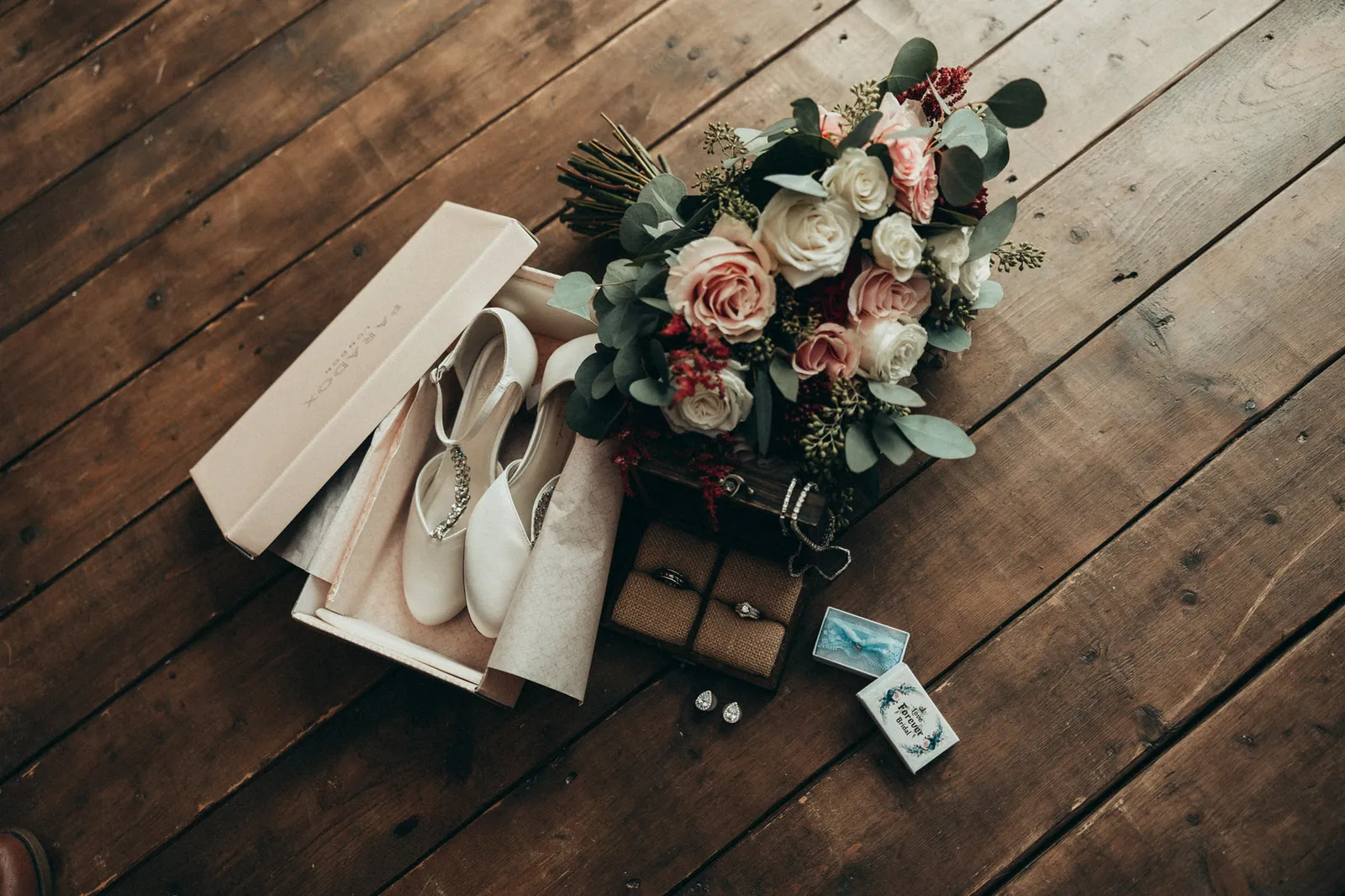 Bridal shoes, a bouquet of blush and red roses, and rings on a wooden floor