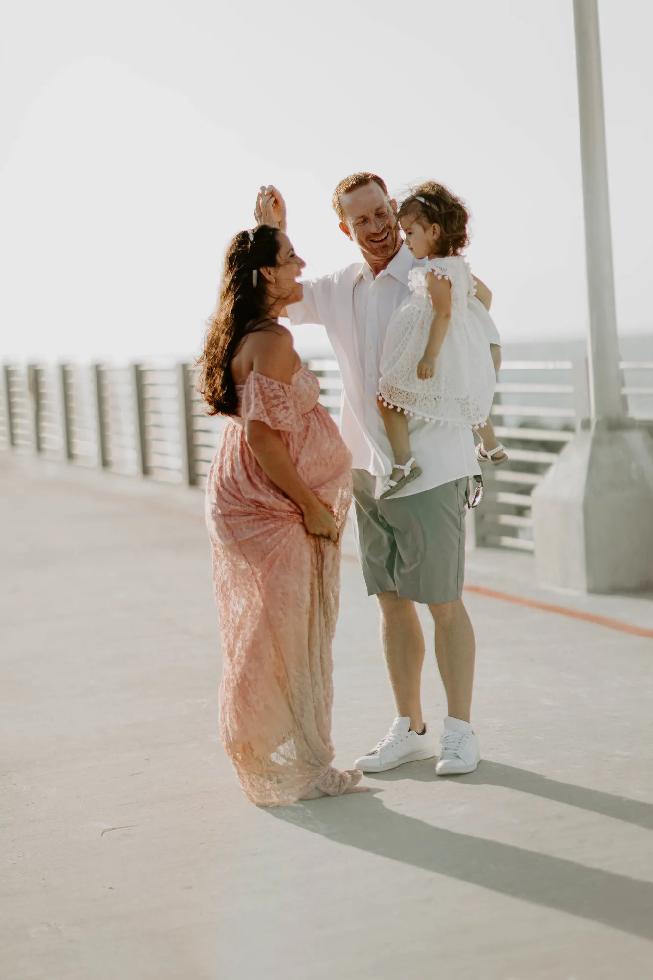 Father holding their daughter as the expectant mother laughs beside them on the pier