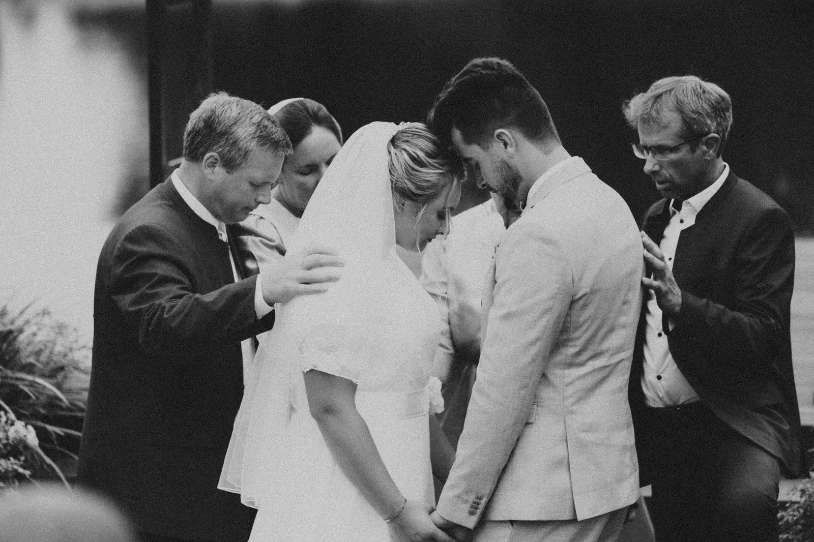 Two men praying over the bride and groom, heads bowed, in black and white