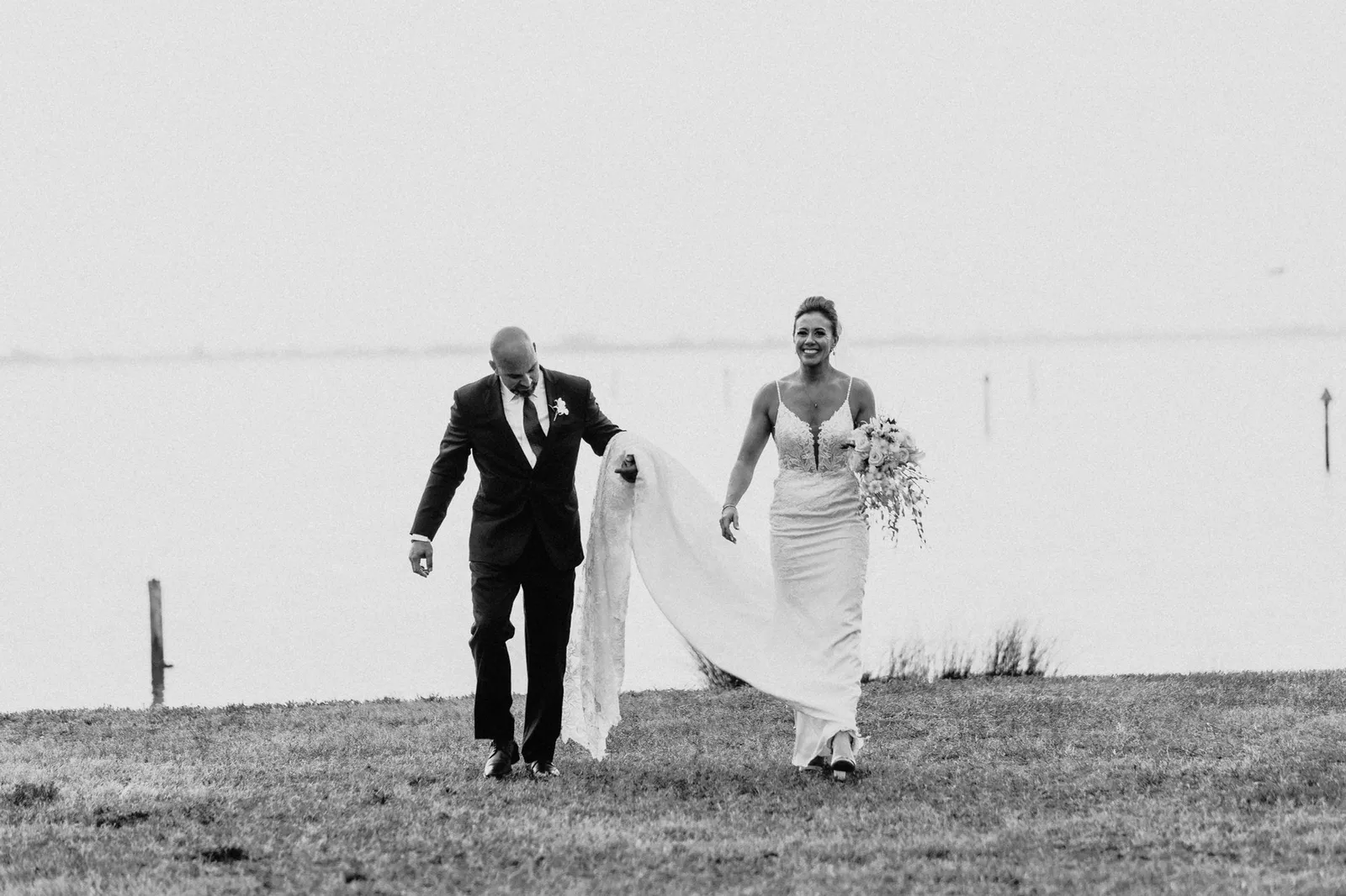 Groom carrying the bride's train as they walk beside the bay, in black and white