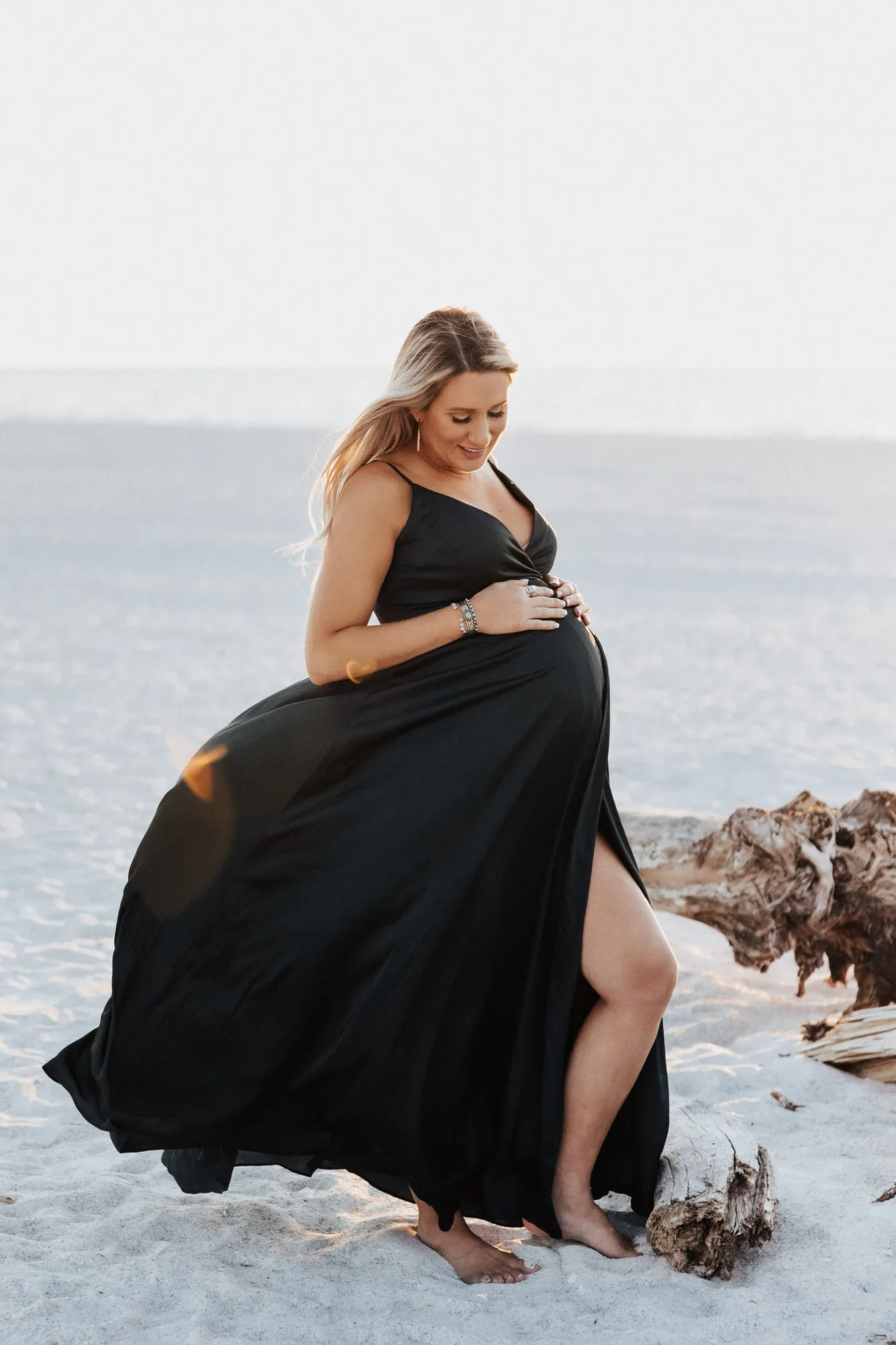 Expectant mother laughing as her dark gown billows on the white sand