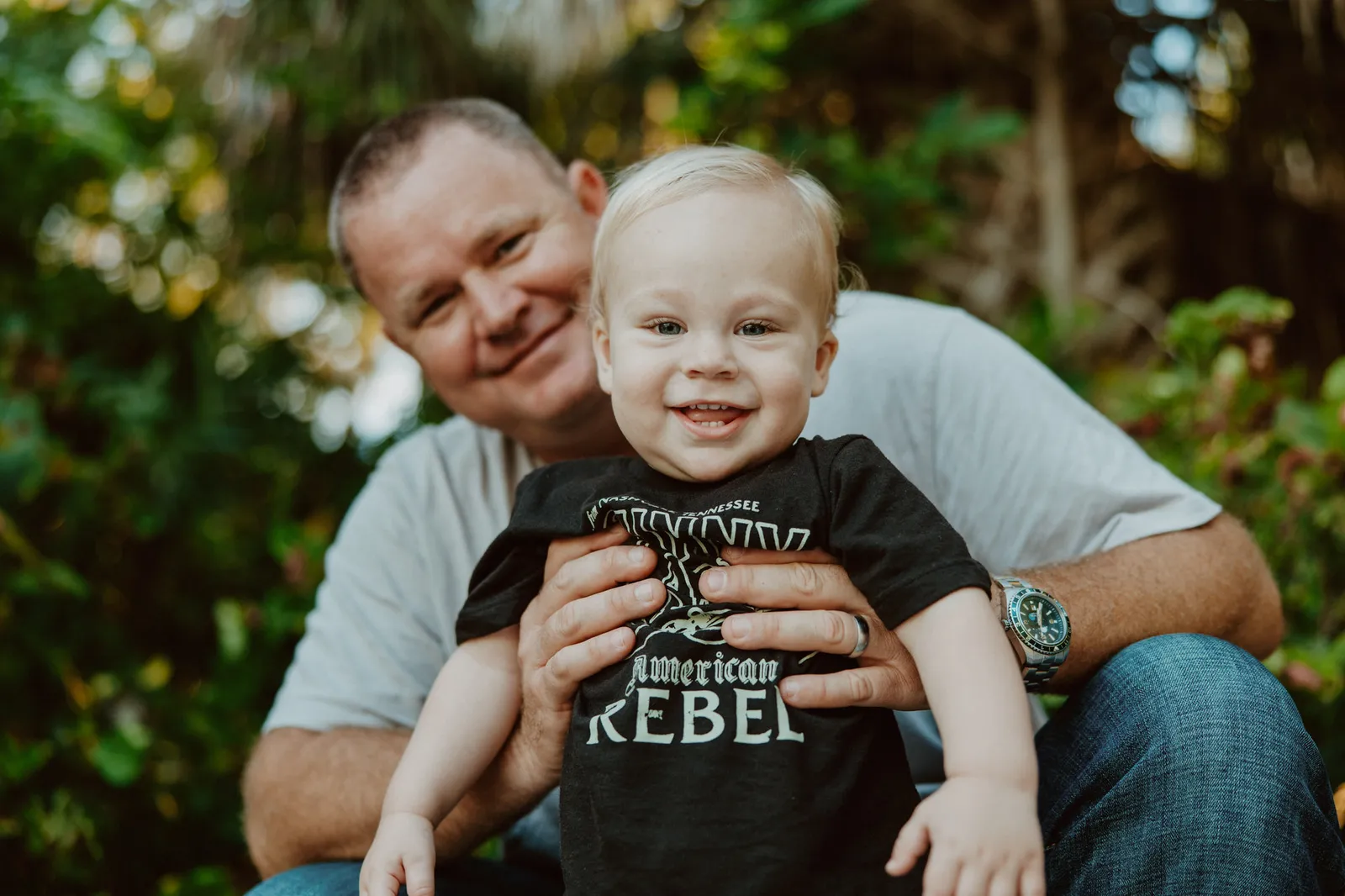 Toddler grinning in a black T-shirt, held up by his father