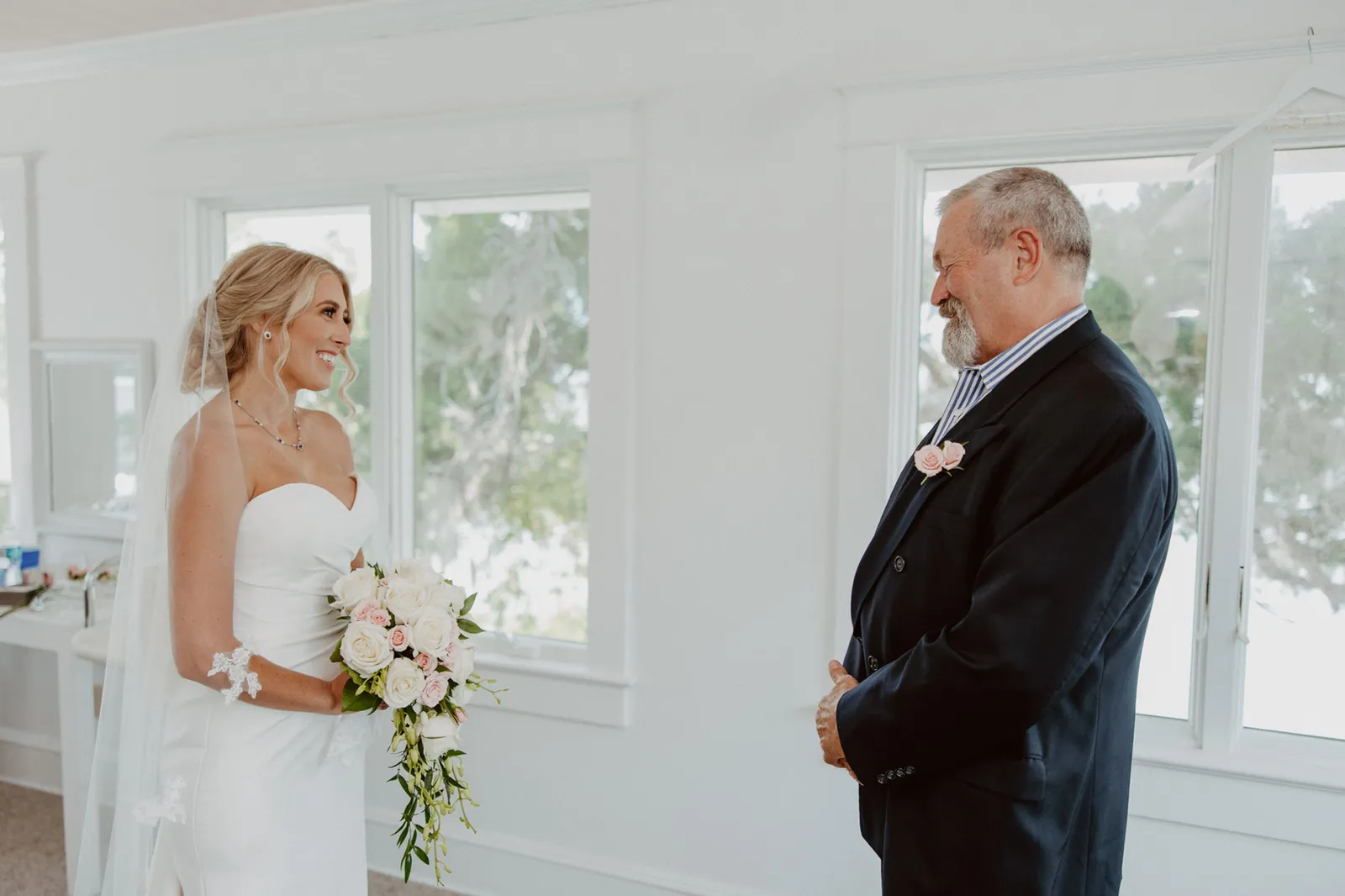 Bride and a man in a dark jacket smiling at each other by the window