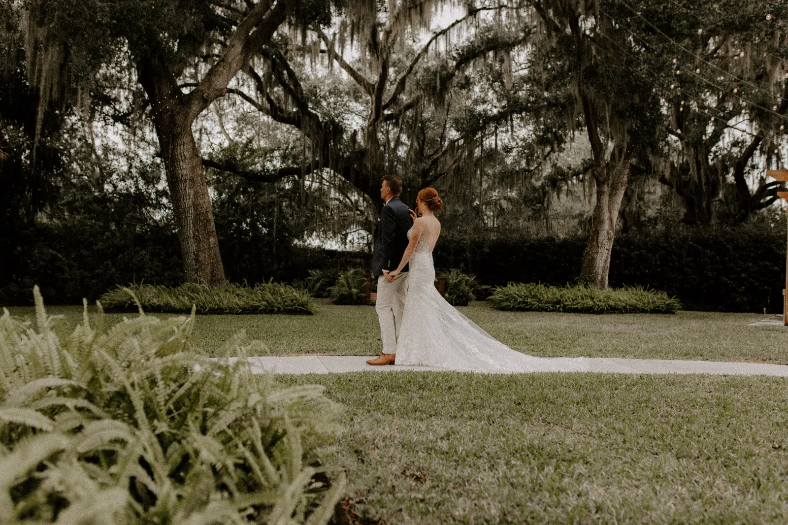 Bride and groom standing together under moss-draped oaks, seen from across the lawn