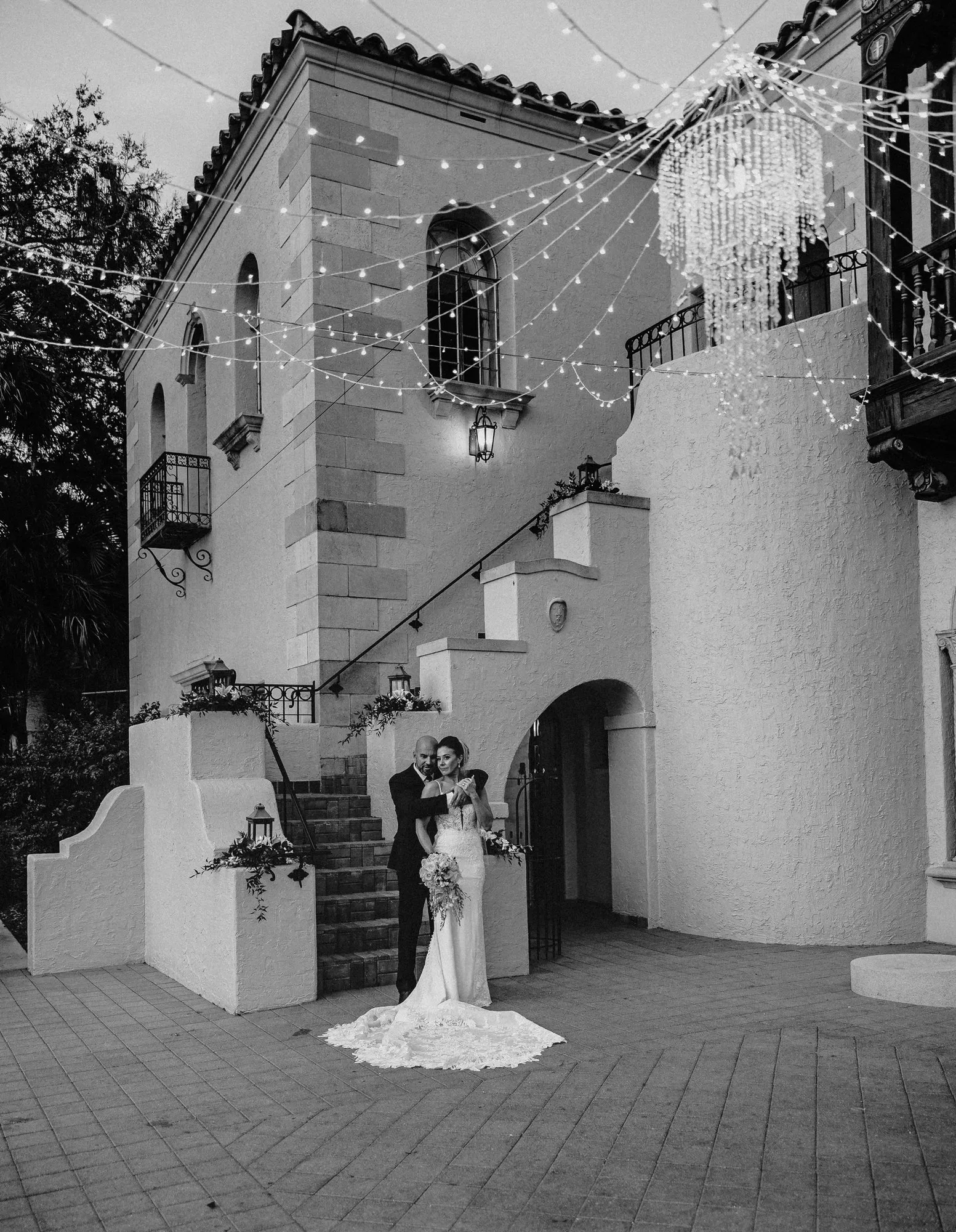 Bride and groom embracing below the estate's tower and string lights, in black and white
