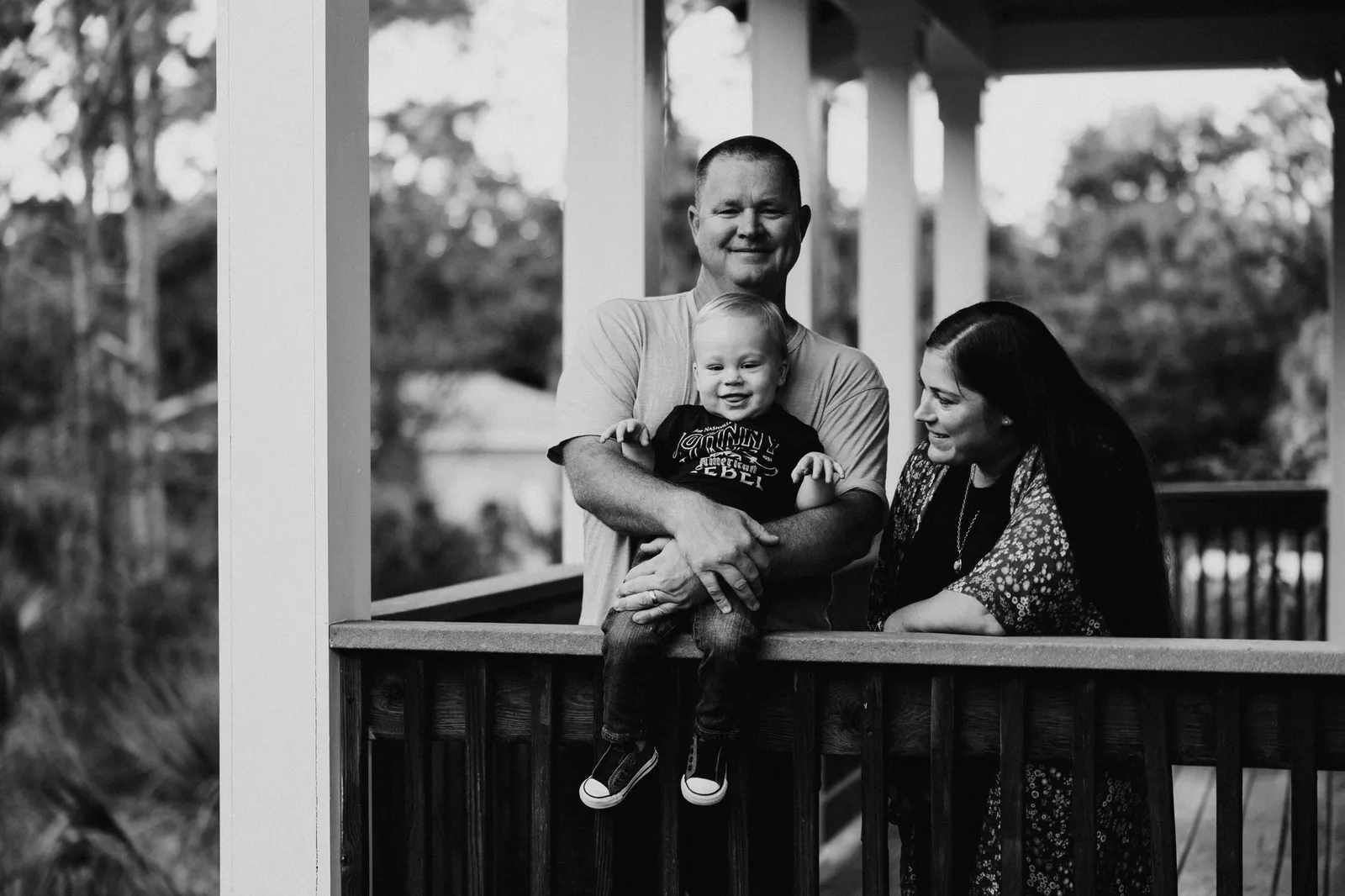 Father holding his son on a porch railing while the mother looks on, in black and white