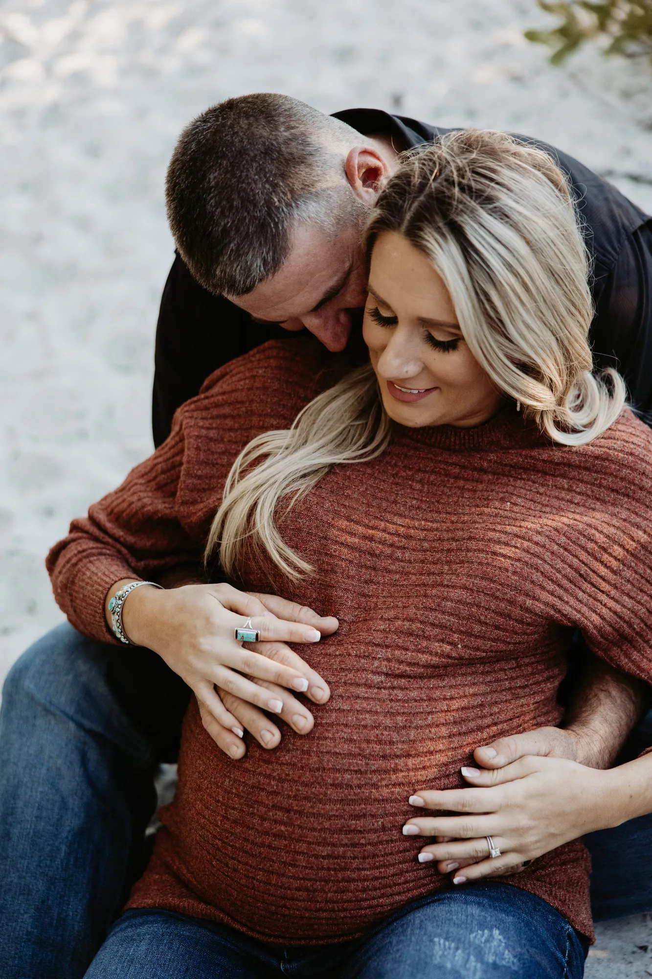 Father-to-be sitting behind her on the sand with his arms around her bump