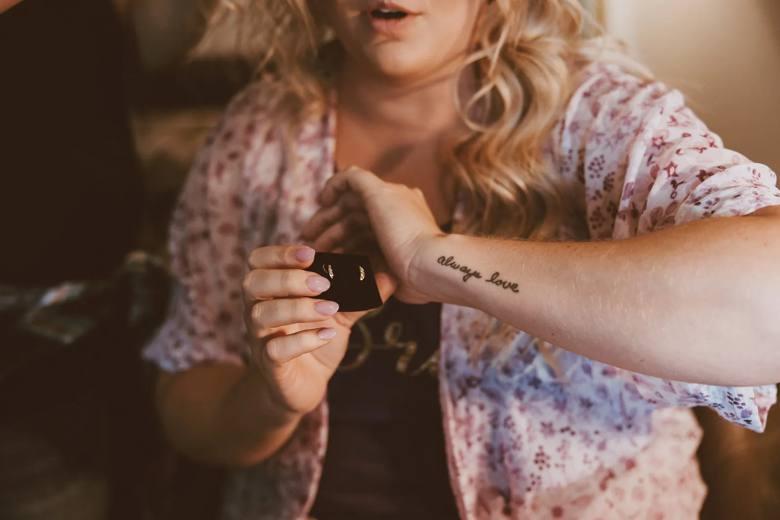 Bride in a floral robe opening a small gift box, a script tattoo on her wrist