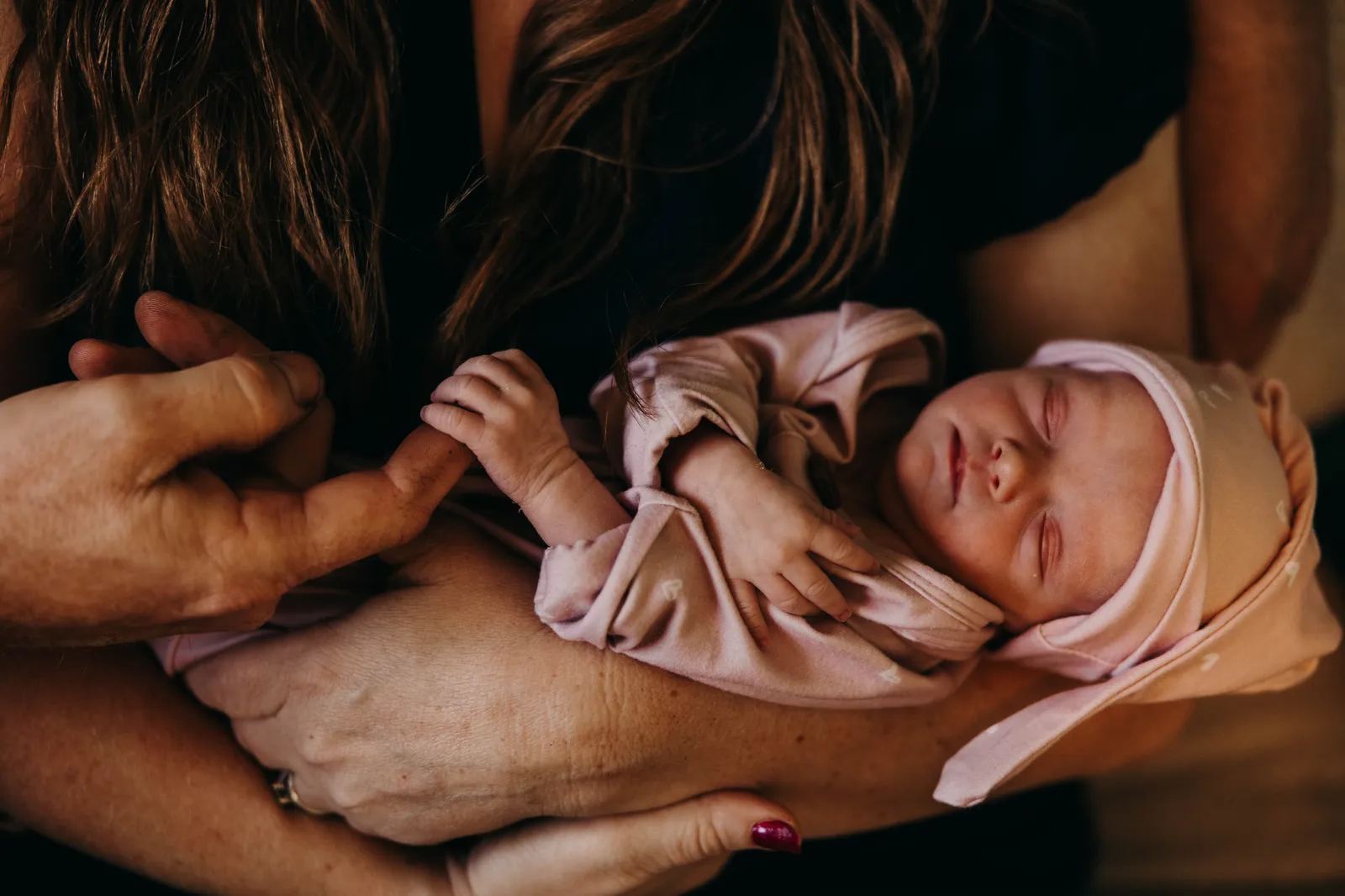 Parents' hands cradling their sleeping newborn in a pink cap