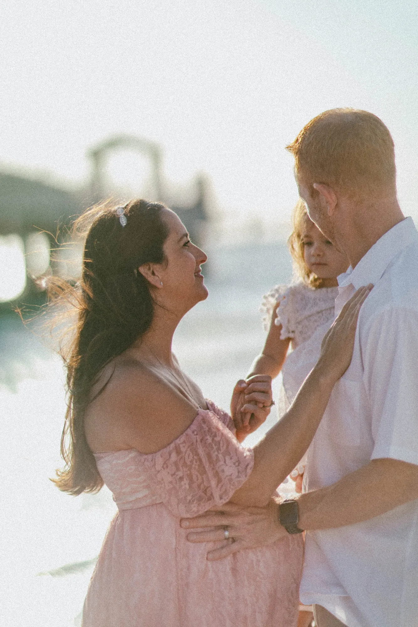 Mother reaching up to her toddler in her husband's arms, backlit by the sun