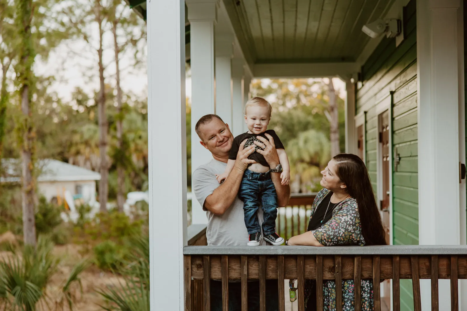 Toddler standing on a porch railing in his father's hands as the mother watches