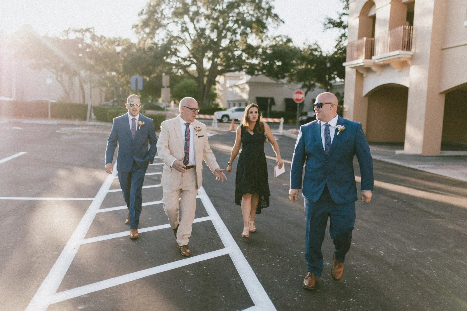 Three men in suits and a woman in a black dress walking across a parking lot in the sun