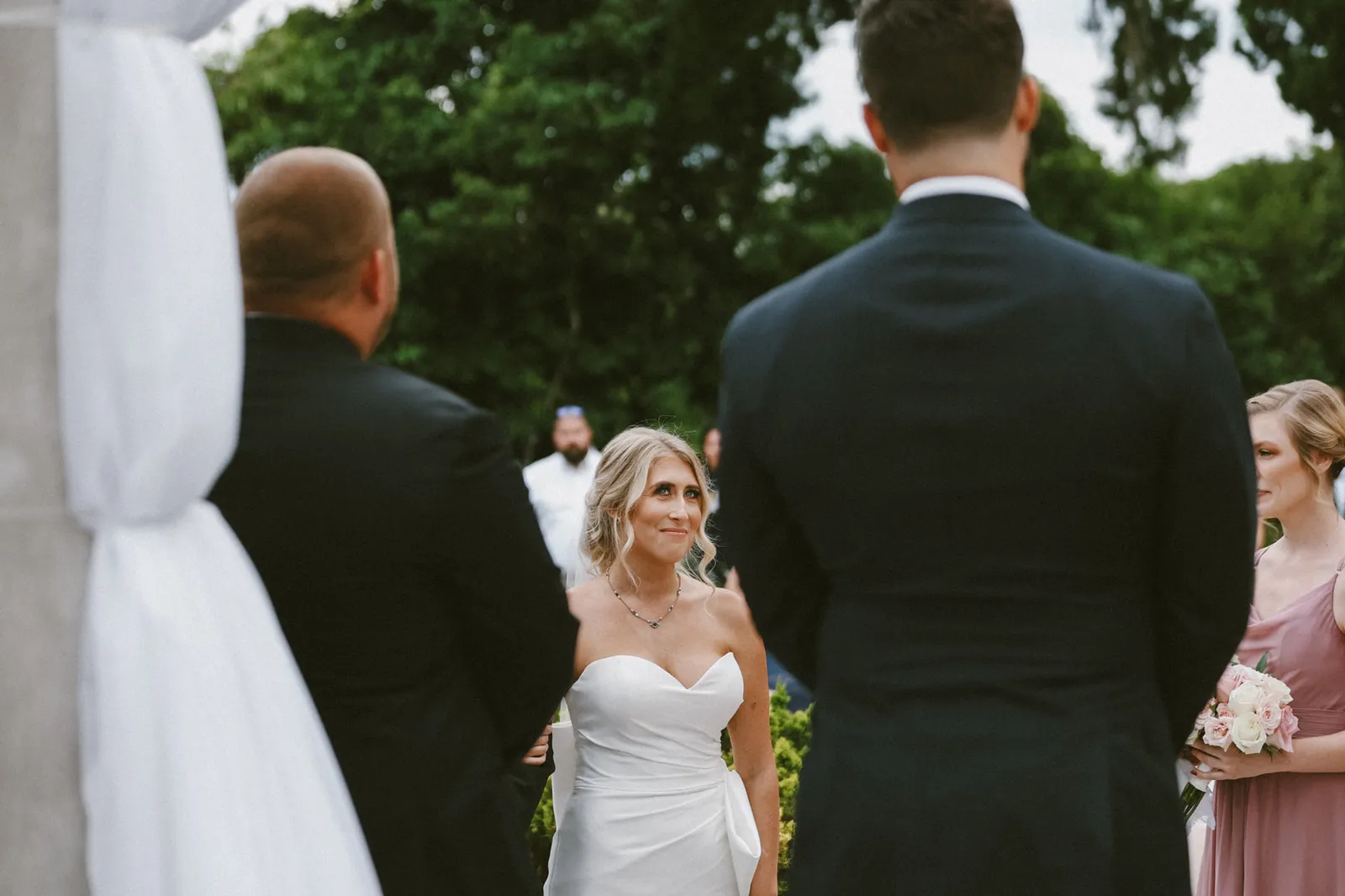 Bride smiling as she approaches the groom, seen between the men waiting at the altar