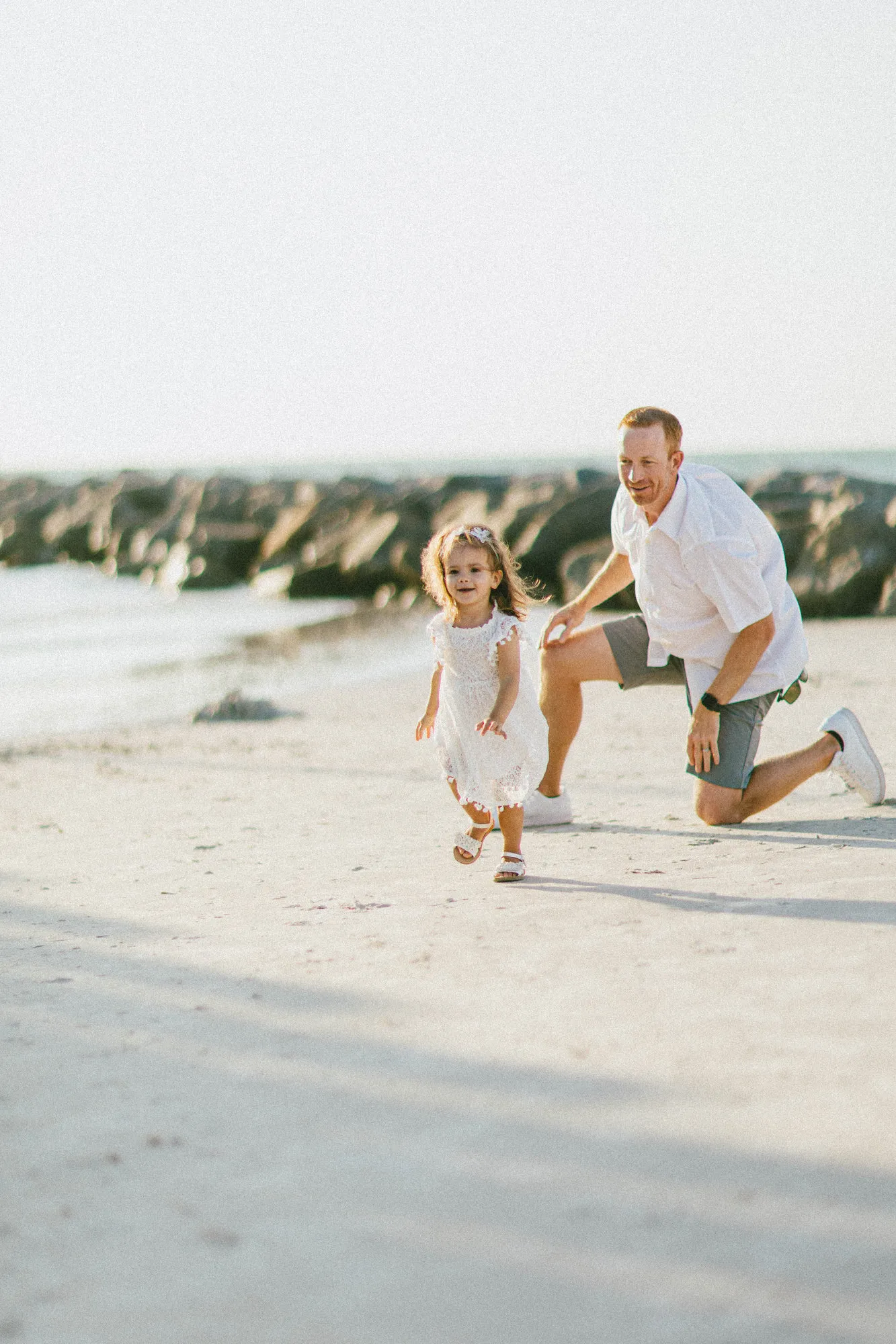 Toddler running toward the camera as her father kneels behind her
