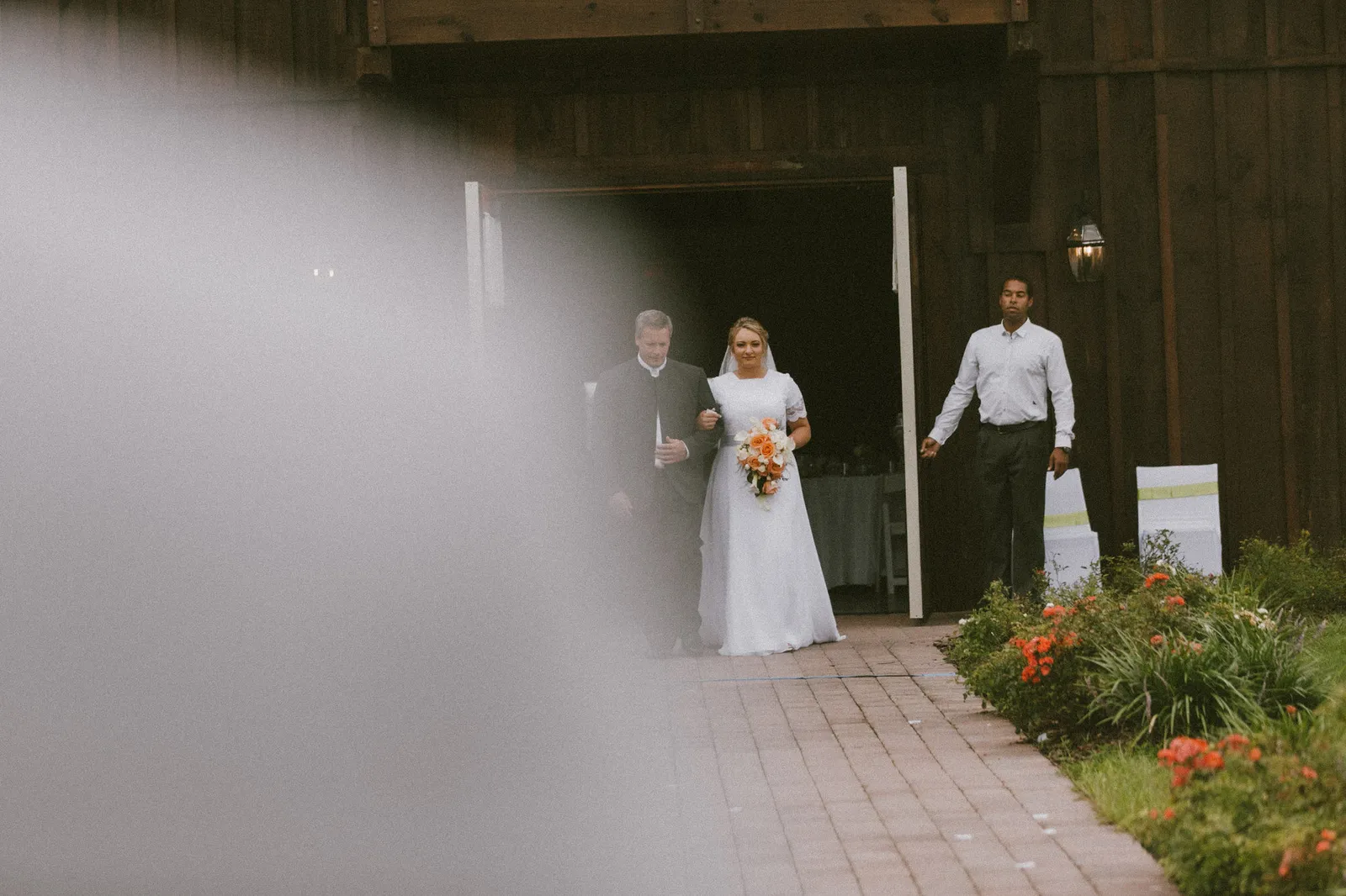 Bride arriving at the top of the brick aisle on a man's arm, seen past a guest's shoulder