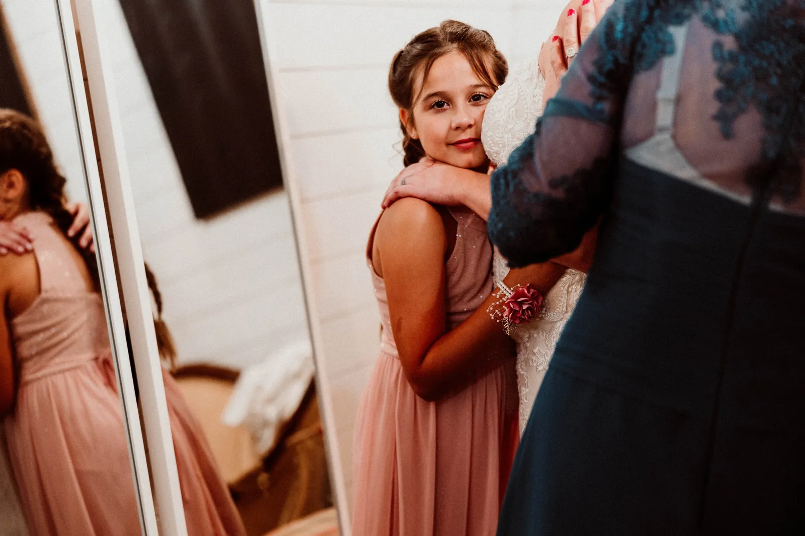 Girl in a pink dress hugging the bride's waist beside a mirror