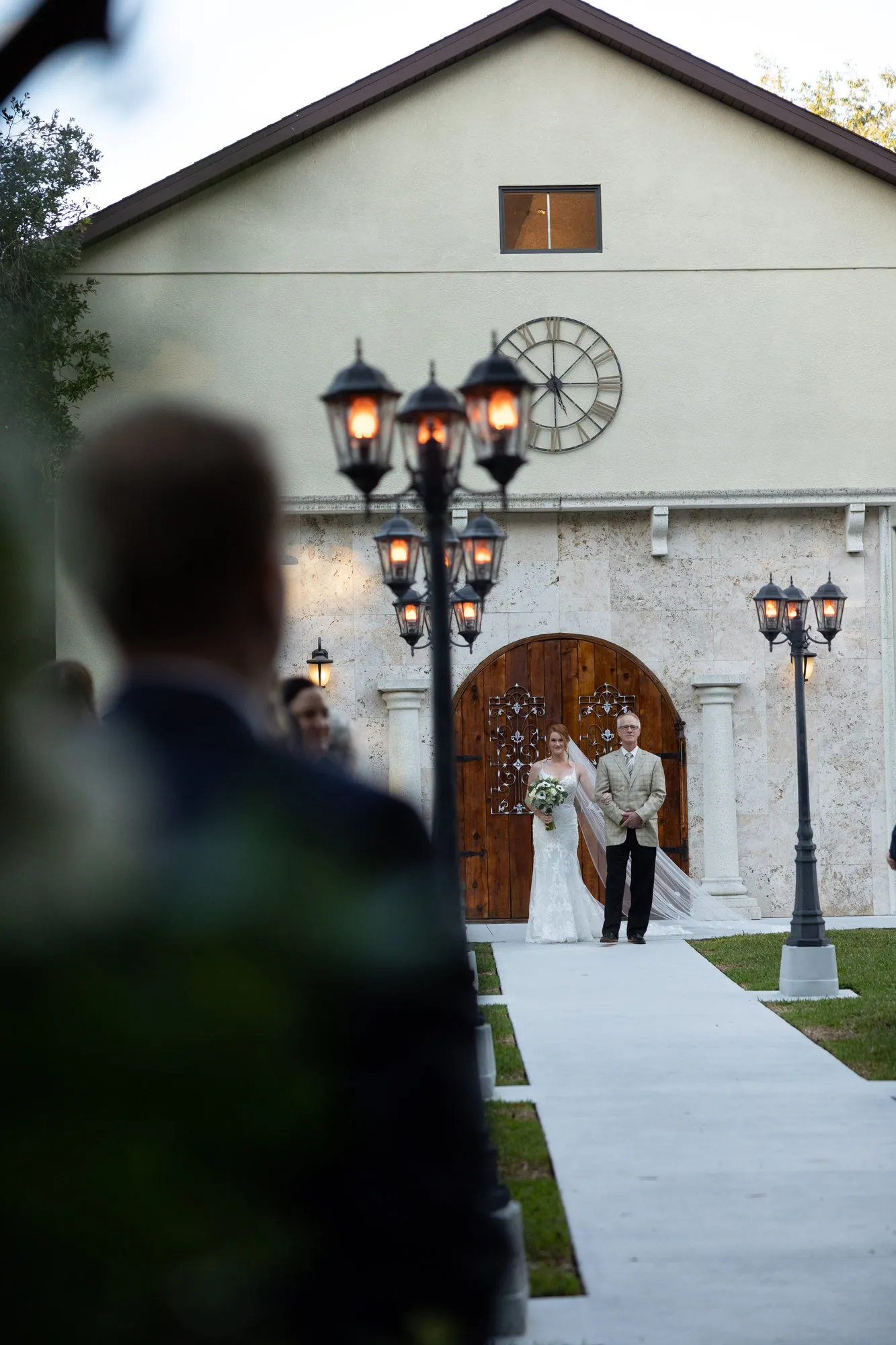 Bride and her escort stepping out of arched wooden doors, seen past the waiting groom
