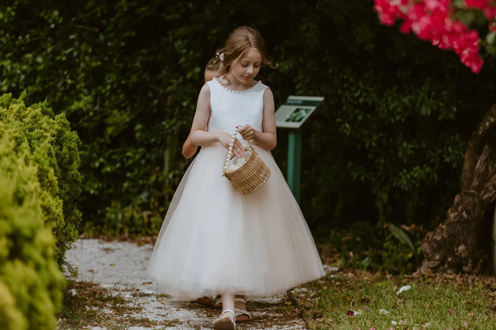 Flower girl in a white dress carrying a basket along a garden path