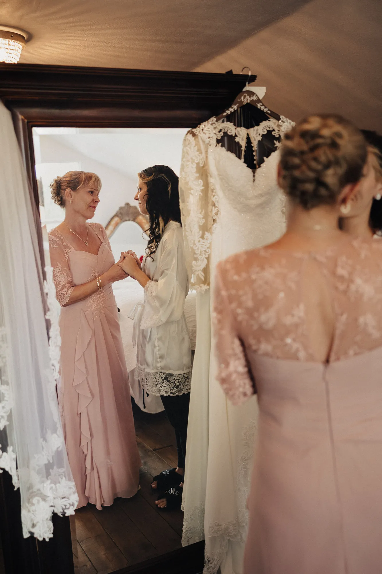 Bride in a robe holding hands with a woman in a blush dress beside her hanging gown