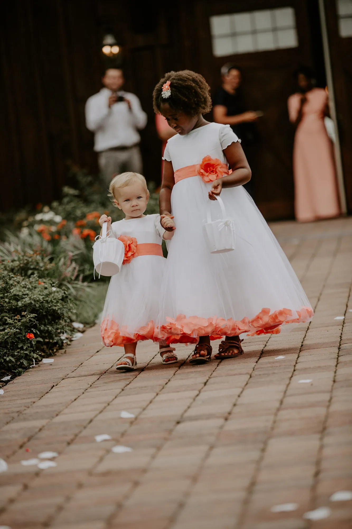 Two small flower girls in white dresses with coral sashes carrying baskets
