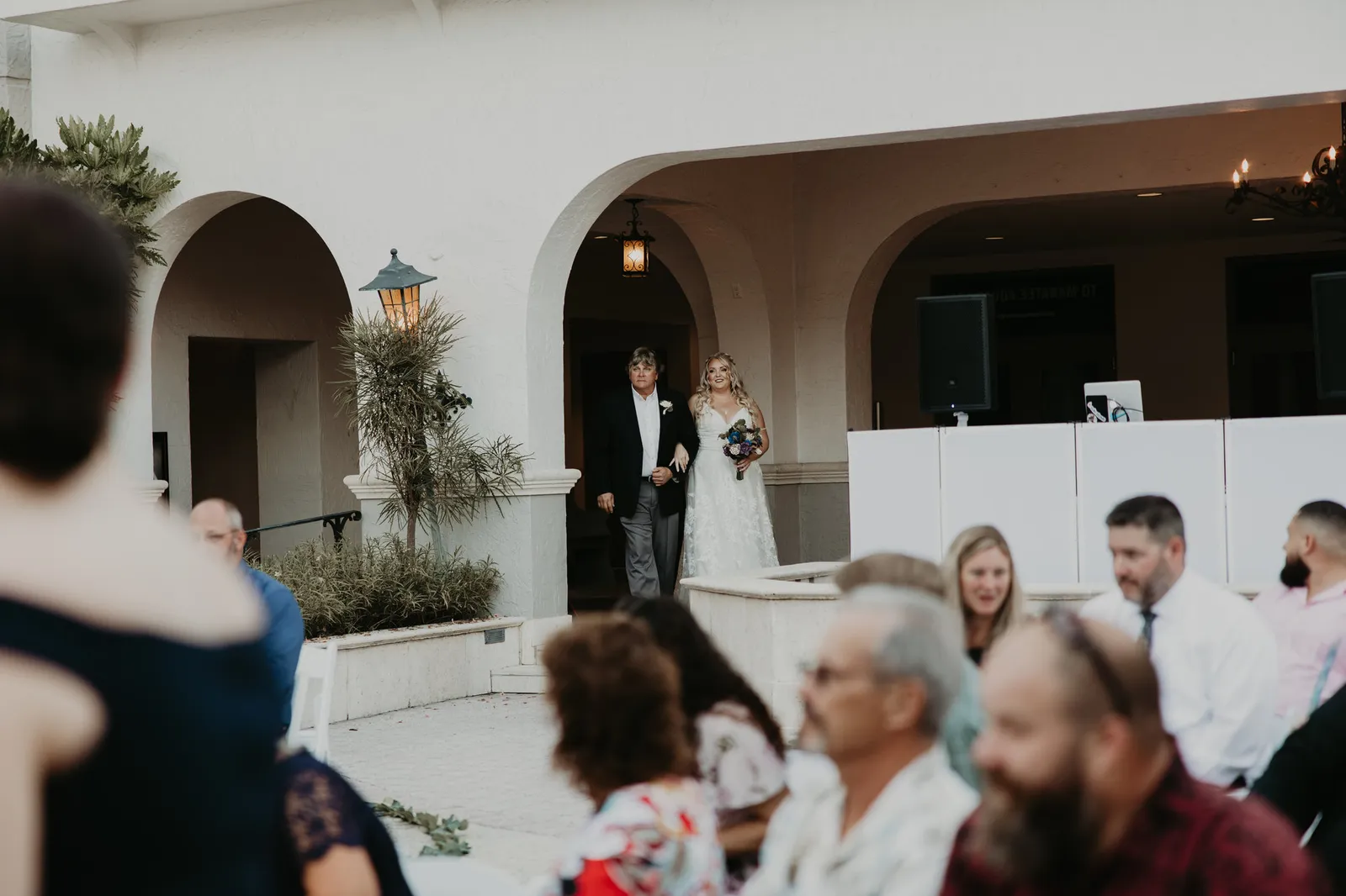 Bride and her escort appearing under the courtyard arches as guests turn to look