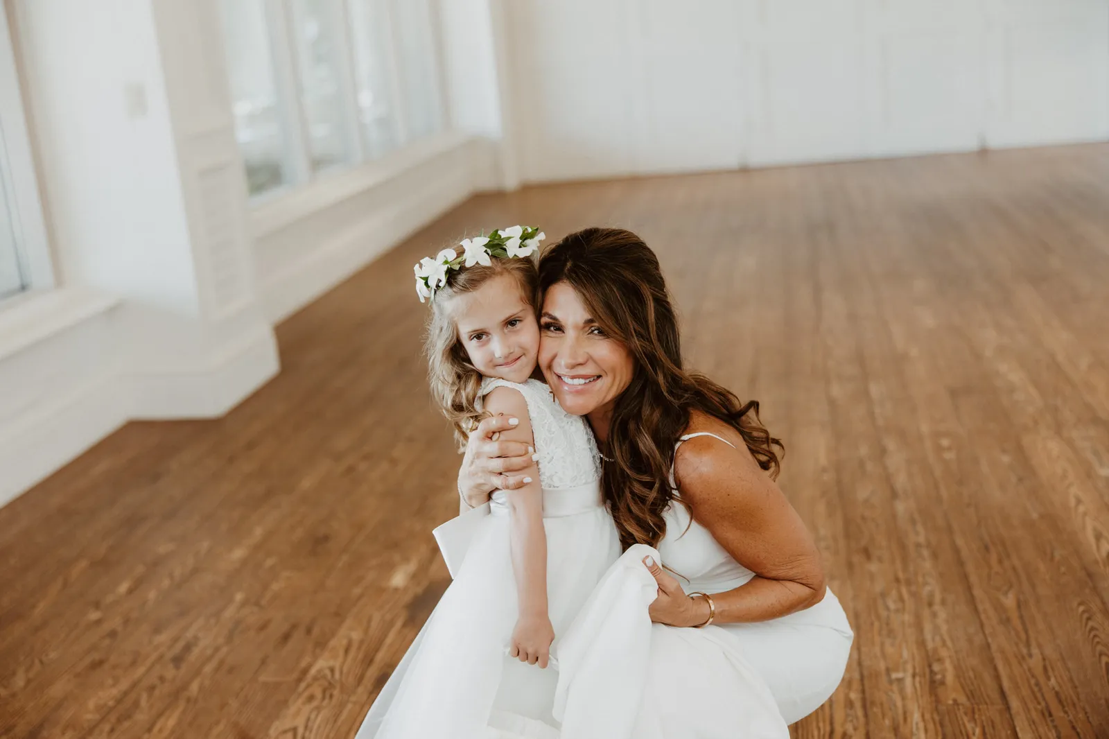 Bride crouching to hug the flower girl, both smiling at the camera