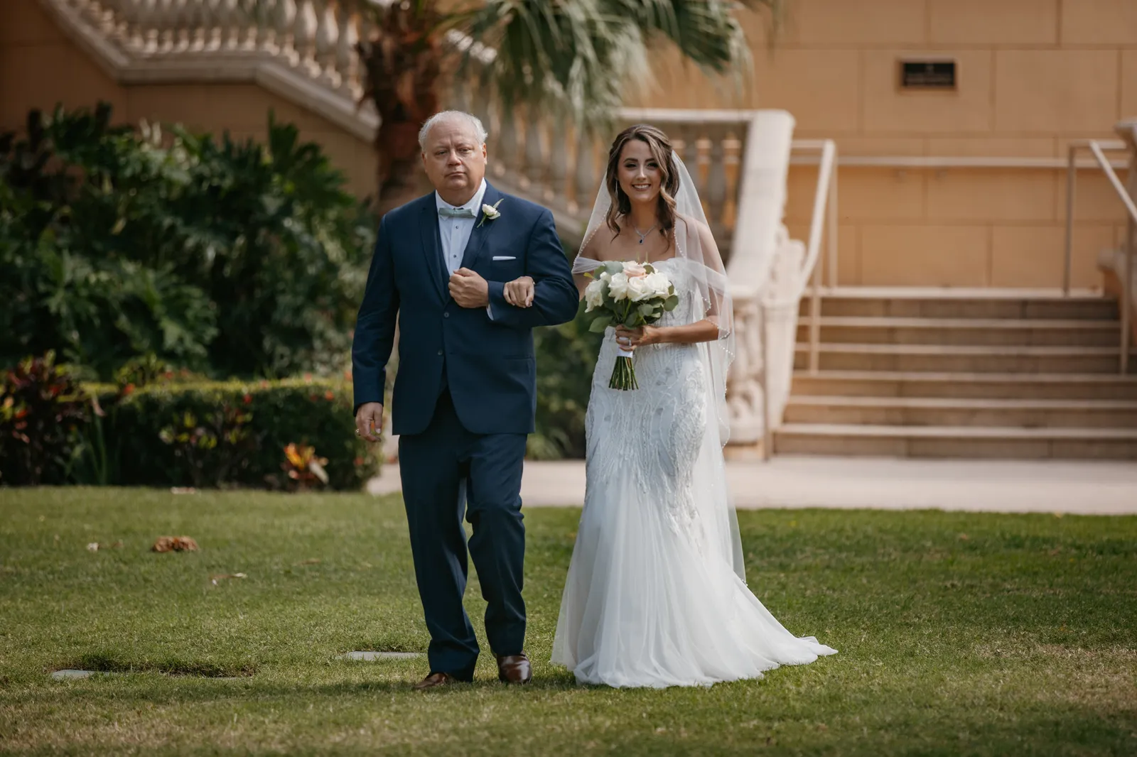 Bride walking across the lawn to the ceremony on the arm of a man in a navy suit